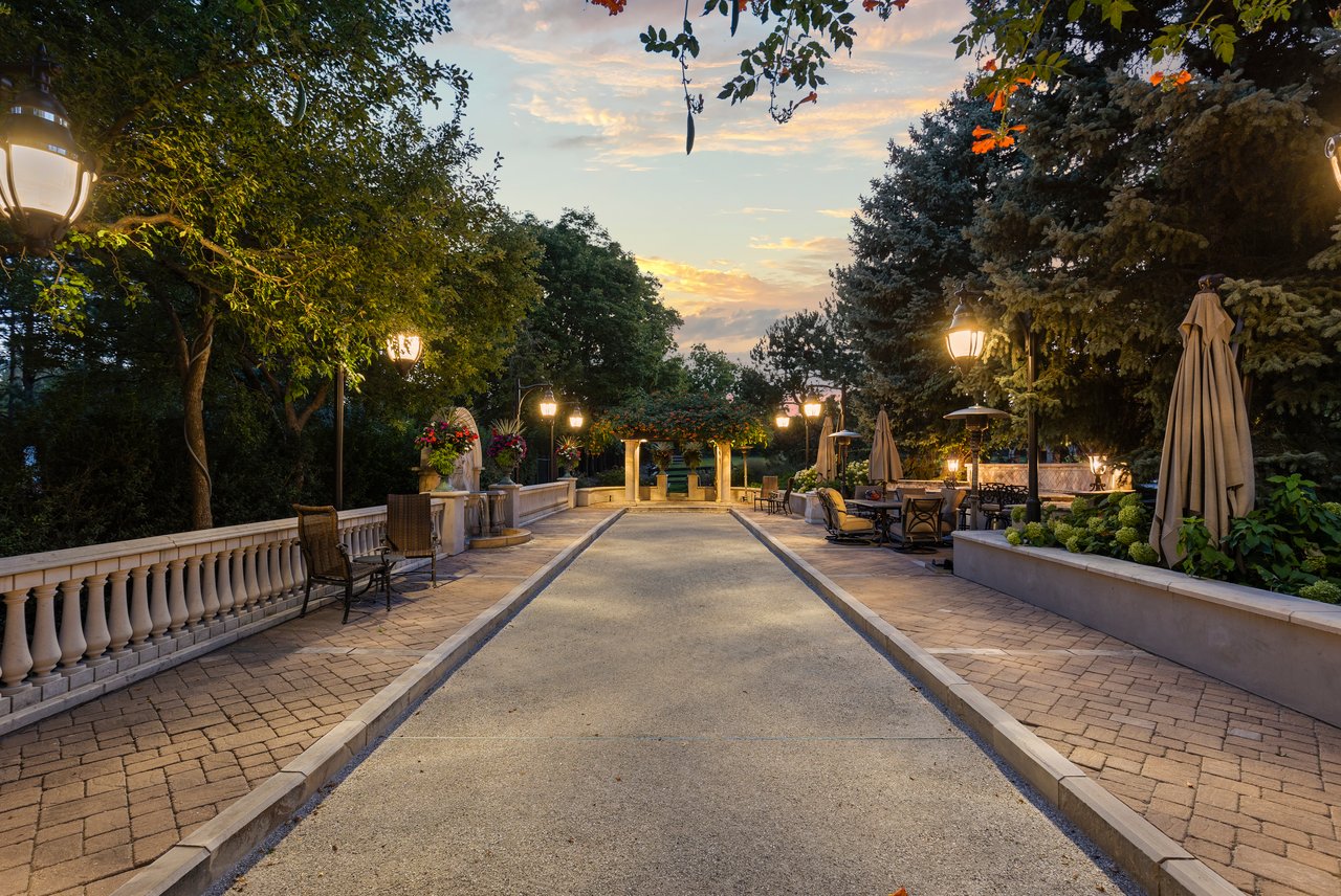 Estate garden patio with a paved walkway, outdoor seating, lush planting, and lanterns glowing at dusk.