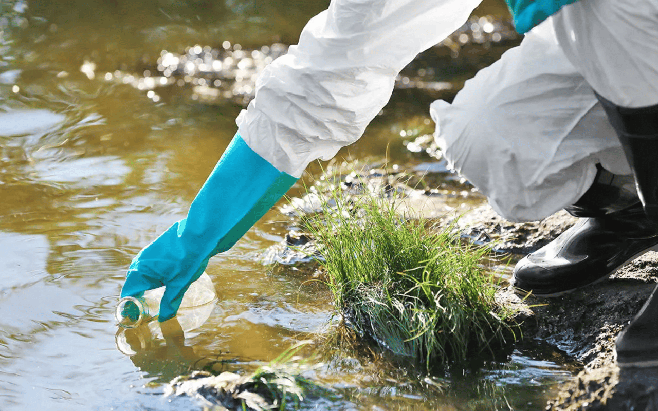 Gloved person in protective clothing retrieves a plastic bottle from a shallow stream.