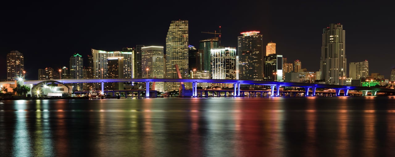 Miami skyline at night, with illuminated high-rises and a blue-lit bridge across the water.
