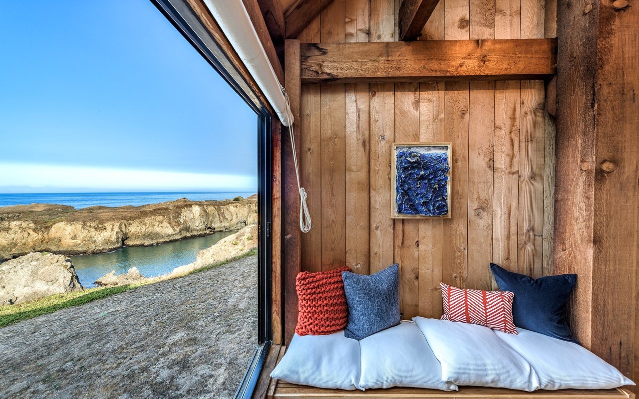 Wood-paneled reading nook with a cushioned window seat overlooking rocky coastline and ocean.