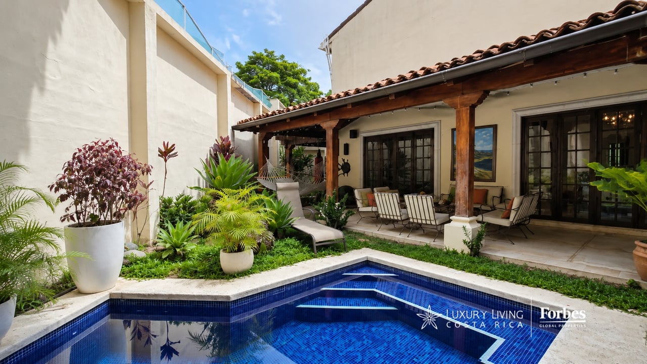 Courtyard swimming pool beside a covered patio with lounge seating and tropical plants.