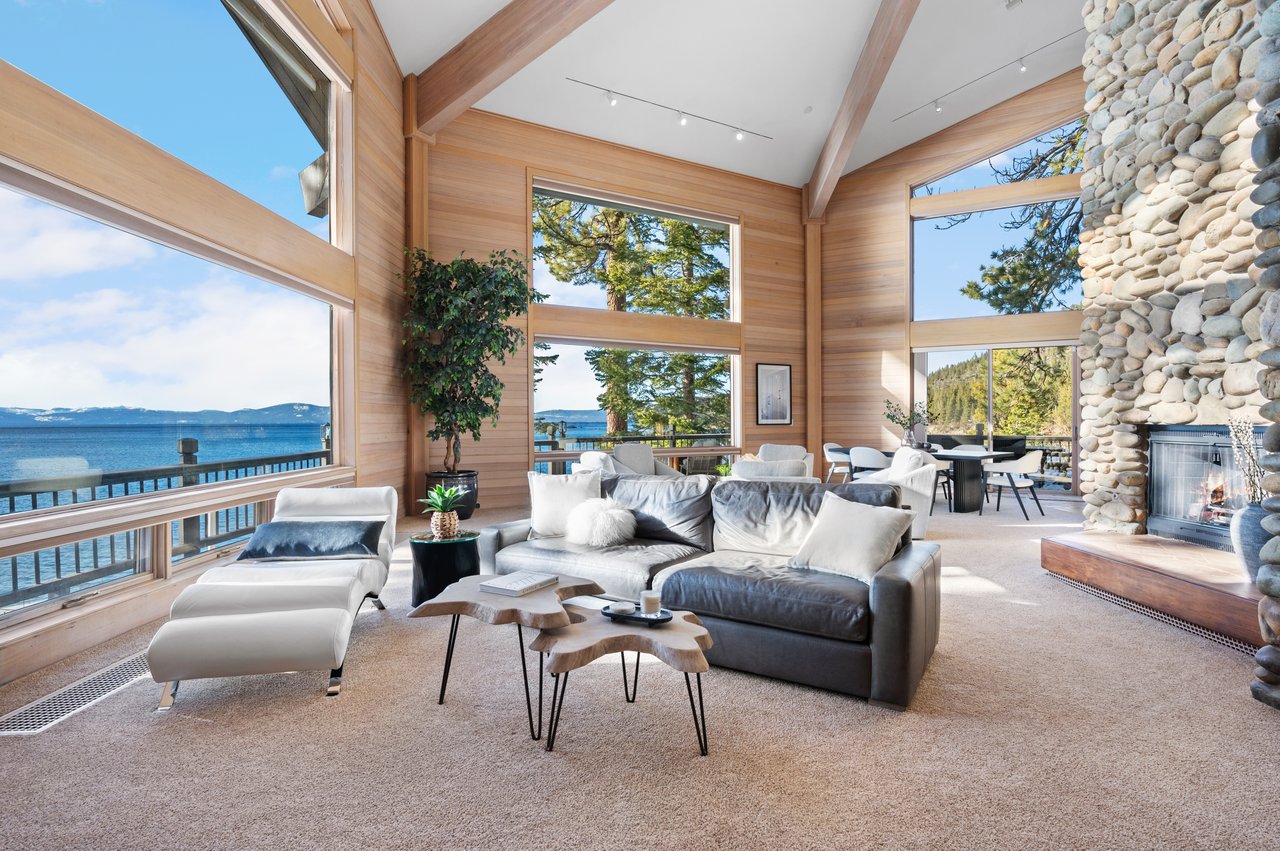 Lakefront living room with vaulted timber ceilings, expansive windows, and a stone fireplace.