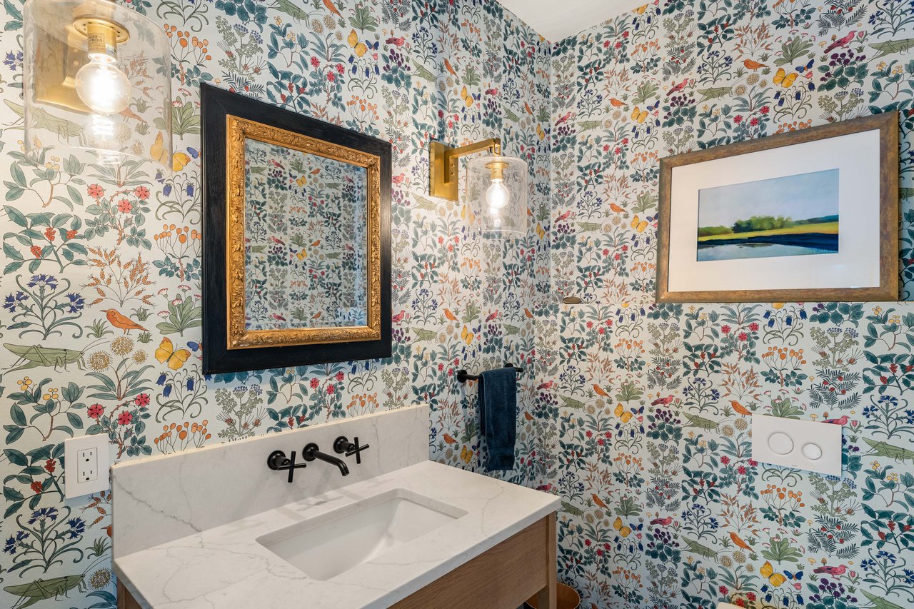 Bathroom vanity with marble countertop, botanical wallpaper, ornate mirror, and brass sconces.