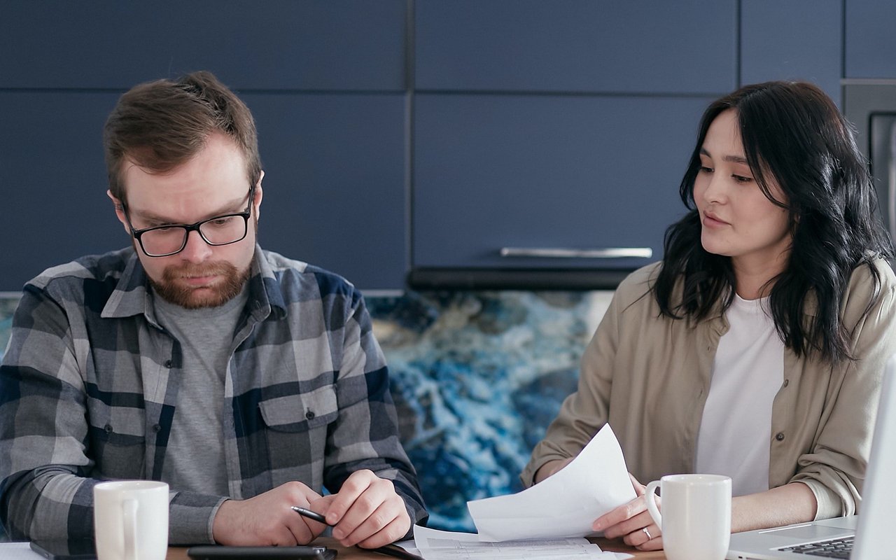 A man and woman review paperwork at a kitchen table, with a laptop and mugs nearby.