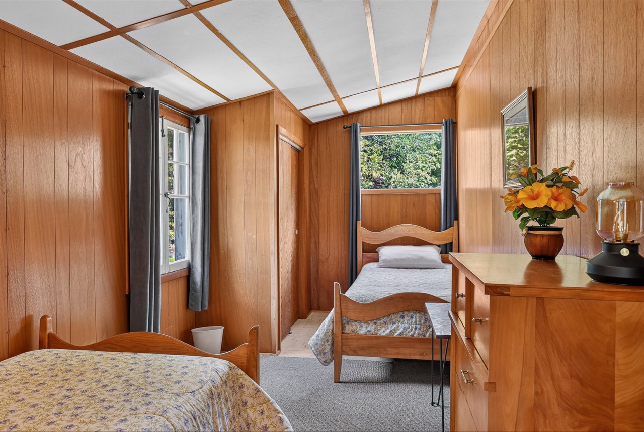 Bedroom with two wooden beds, wood-paneled walls, gray curtains, and a dresser with flowers.