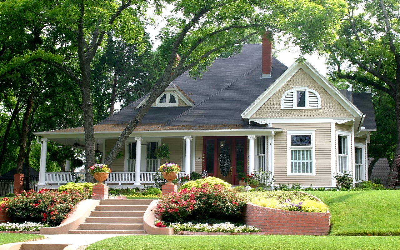 Beige house with a white-columned porch, red front door, flower beds, and broad front steps.