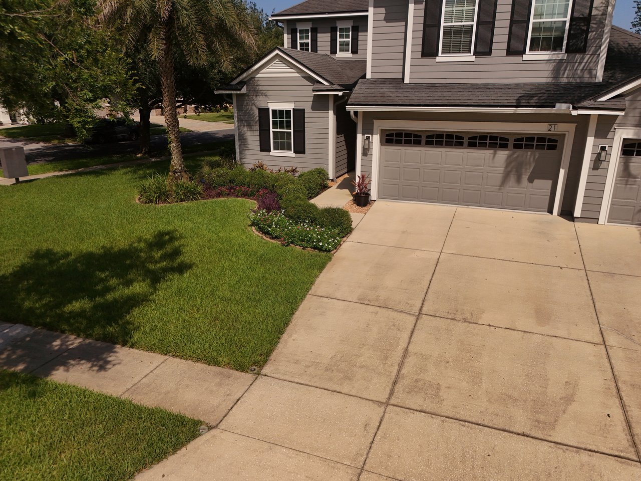 Two-story gray house with black shutters, double garage, landscaped flower beds, and a broad driveway.
