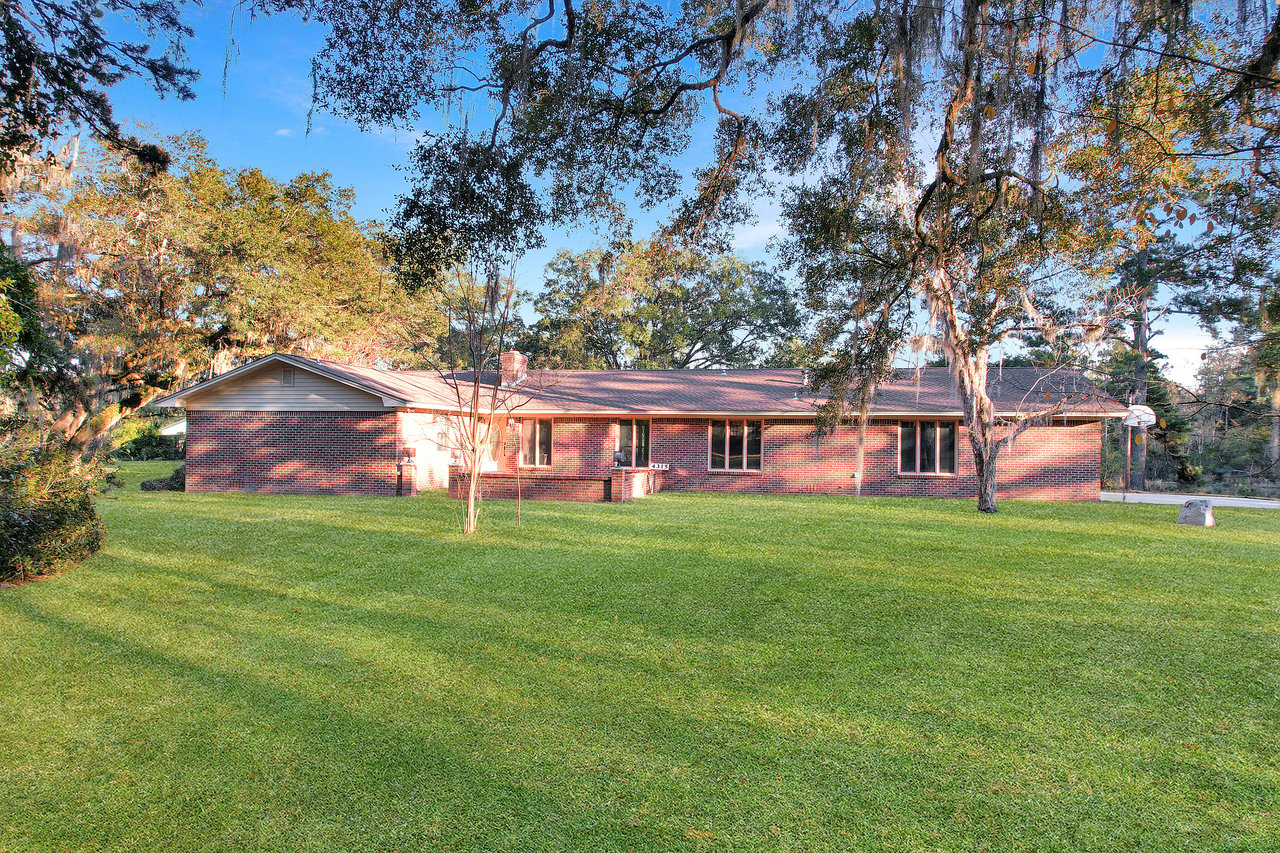 A brick house with multiple windows sits in a lush, green yard under large, leafy trees. The sun casts warm, dappled light, creating a serene atmosphere.