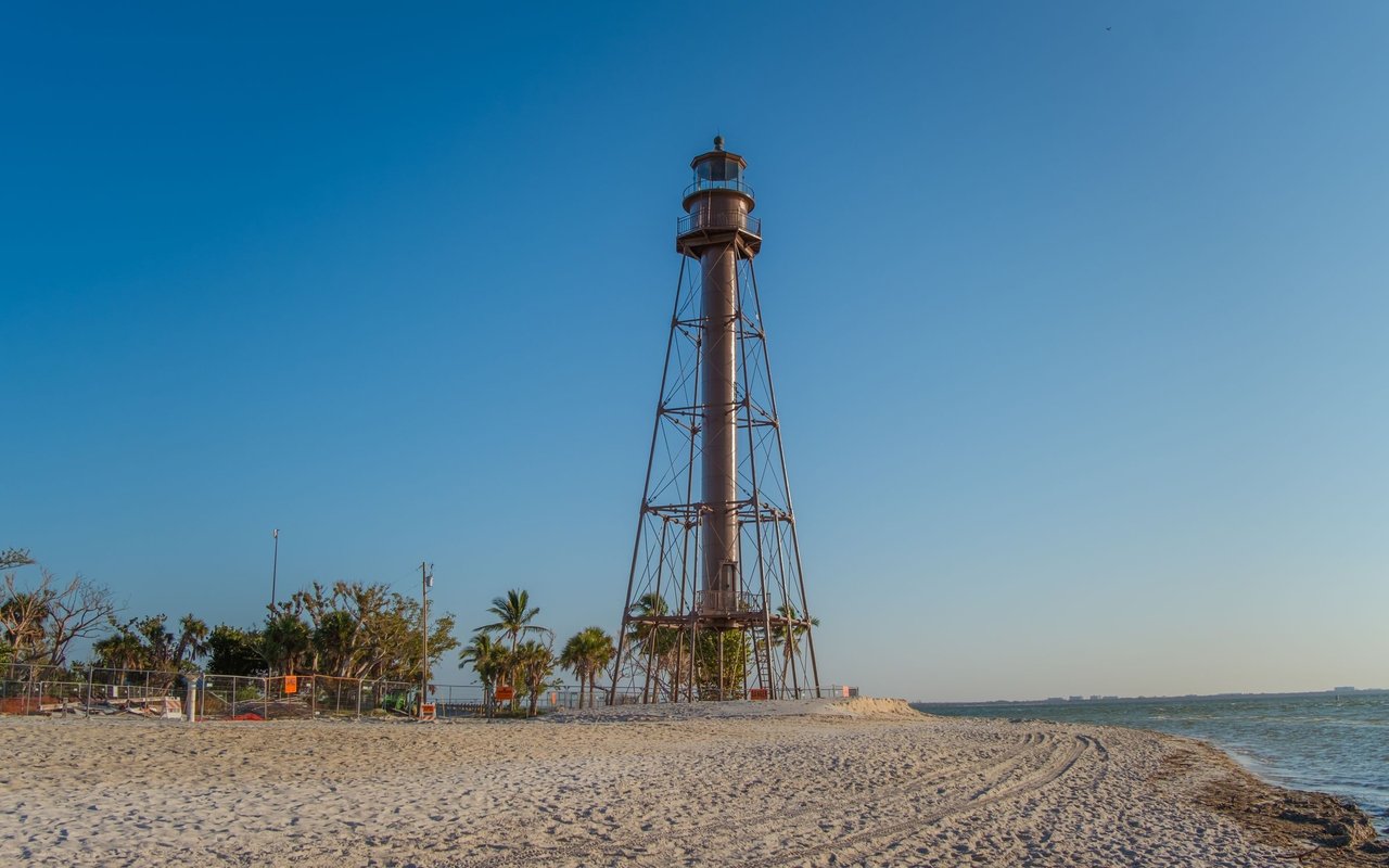 A tall open-frame lighthouse stands on a sandy beach beside coastal water.