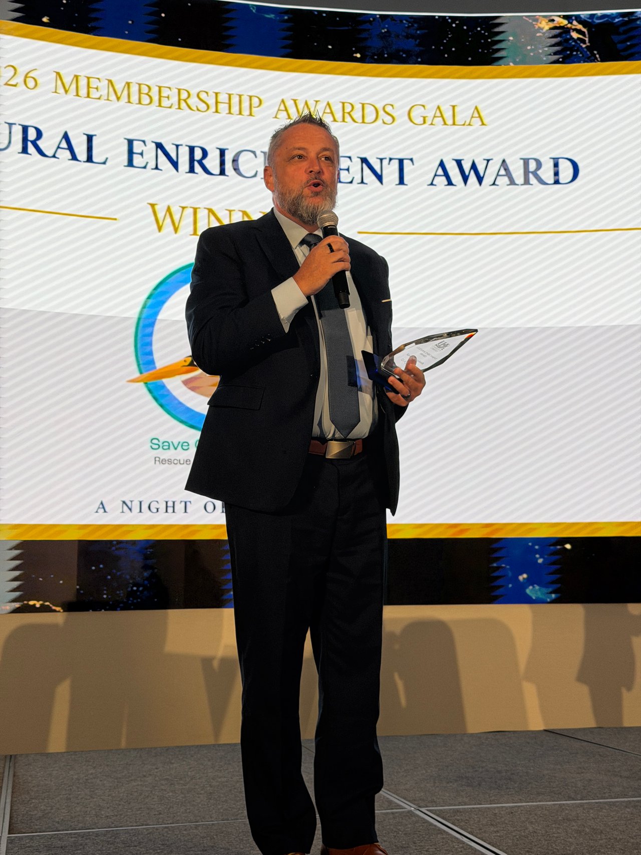 Man in a dark suit speaks into a microphone while holding a plaque at an awards gala.