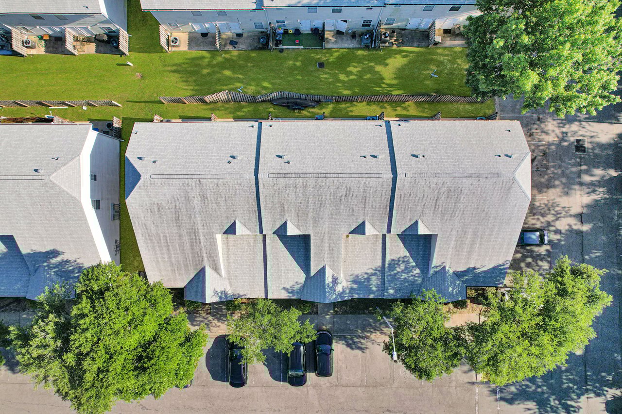 Aerial view of residential roofs with gray shingles. Three trees and three parked cars line the street. Green lawns and fences are visible in the backyard.