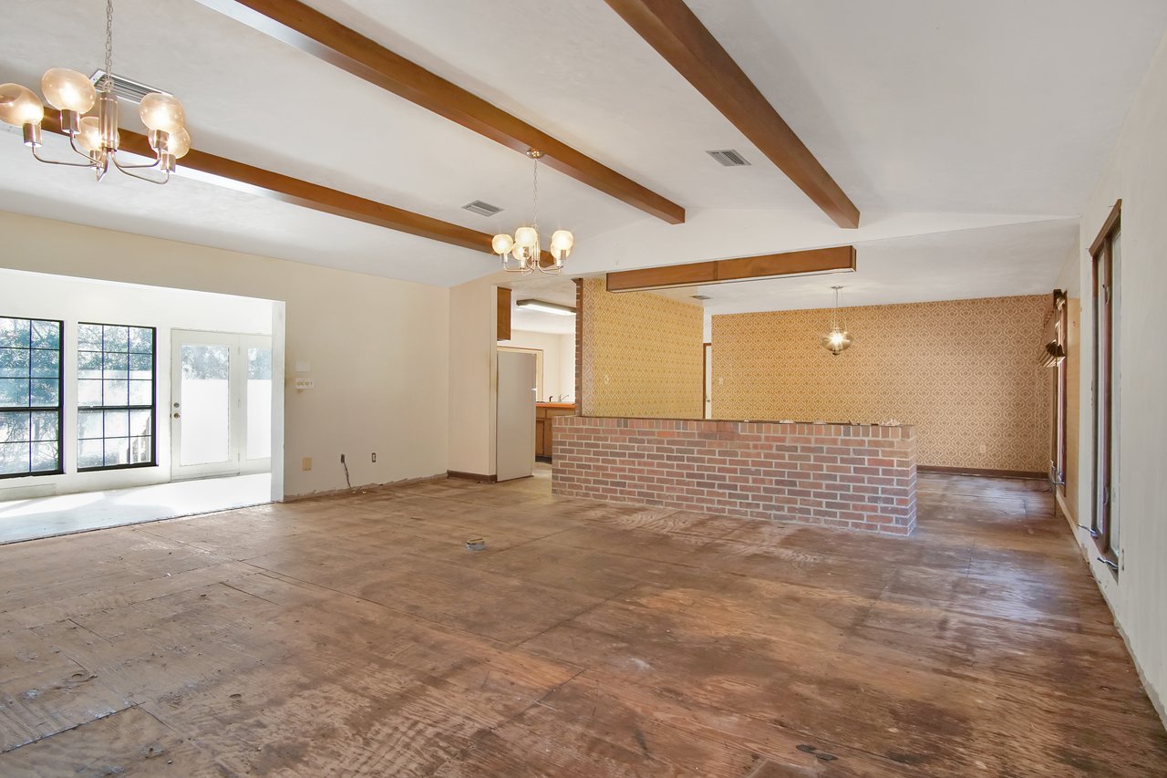 Spacious, unfinished room with exposed wooden floor, brick half-wall, and patterned wallpaper. Sunlit area with windows and double doors, ceiling lights hang from wood beams, creating a rustic feel.