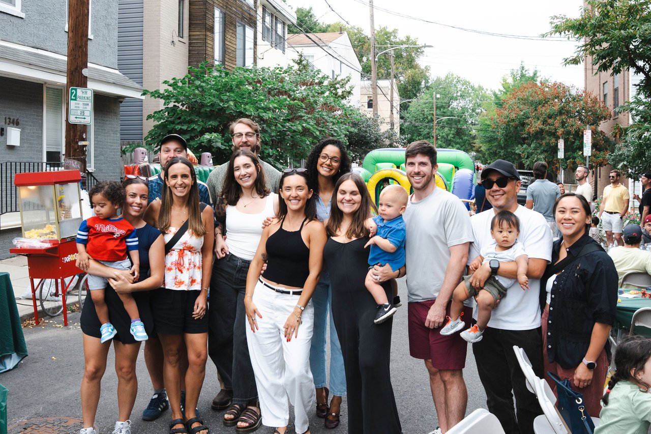 Neighbors pose with children at a block party beside a popcorn cart and inflatable play structure.