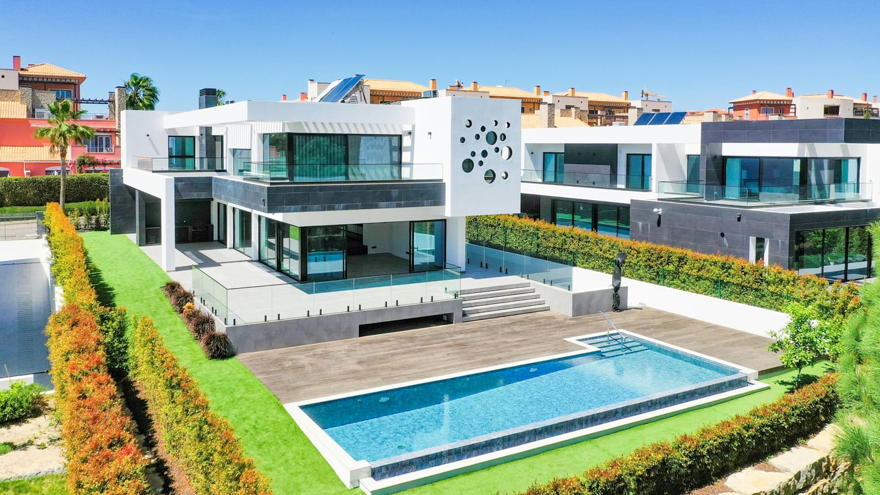 Modern white luxury home with glass balconies, landscaped grounds, and a rectangular swimming pool.