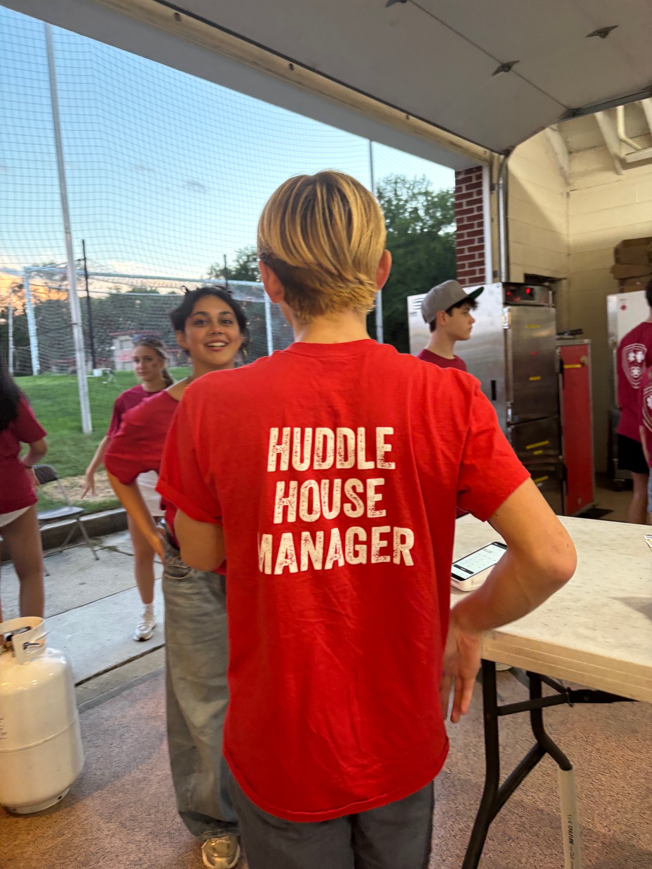 Volunteers gather at a football concession stand; the person in front wears a red Huddle House Manager shirt.