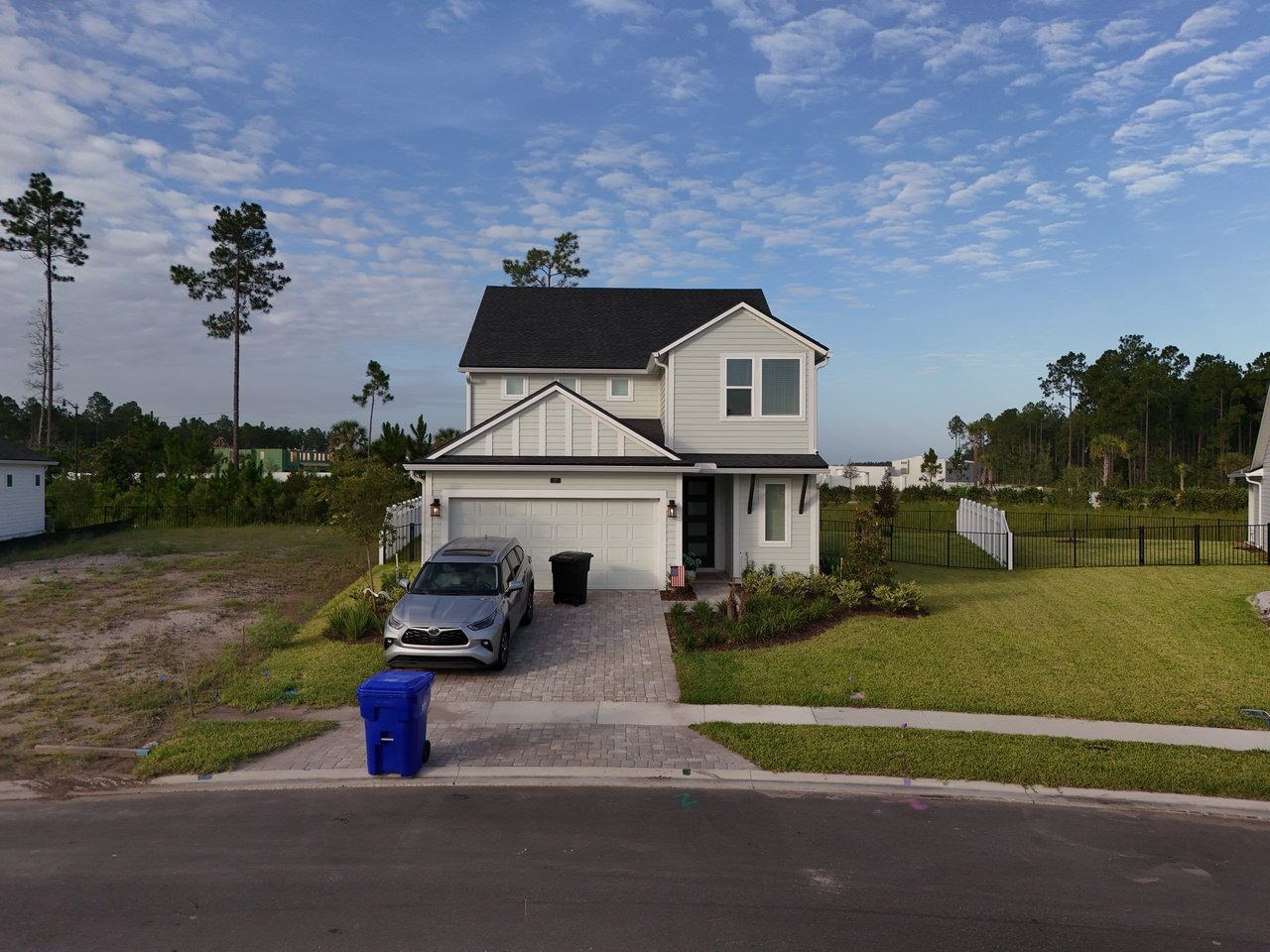 Two-story light gray house with an attached garage, paver driveway, and fenced lawn.