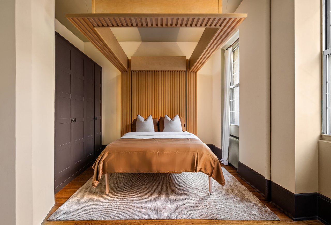Bedroom with a bed framed by a wood-slat canopy, built-in wardrobes, and tall windows.