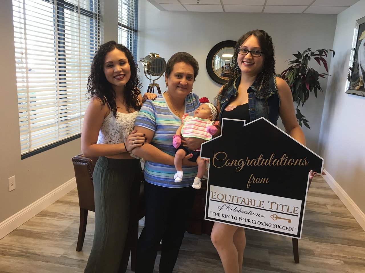Three women pose with a baby beside a congratulatory title-company sign.