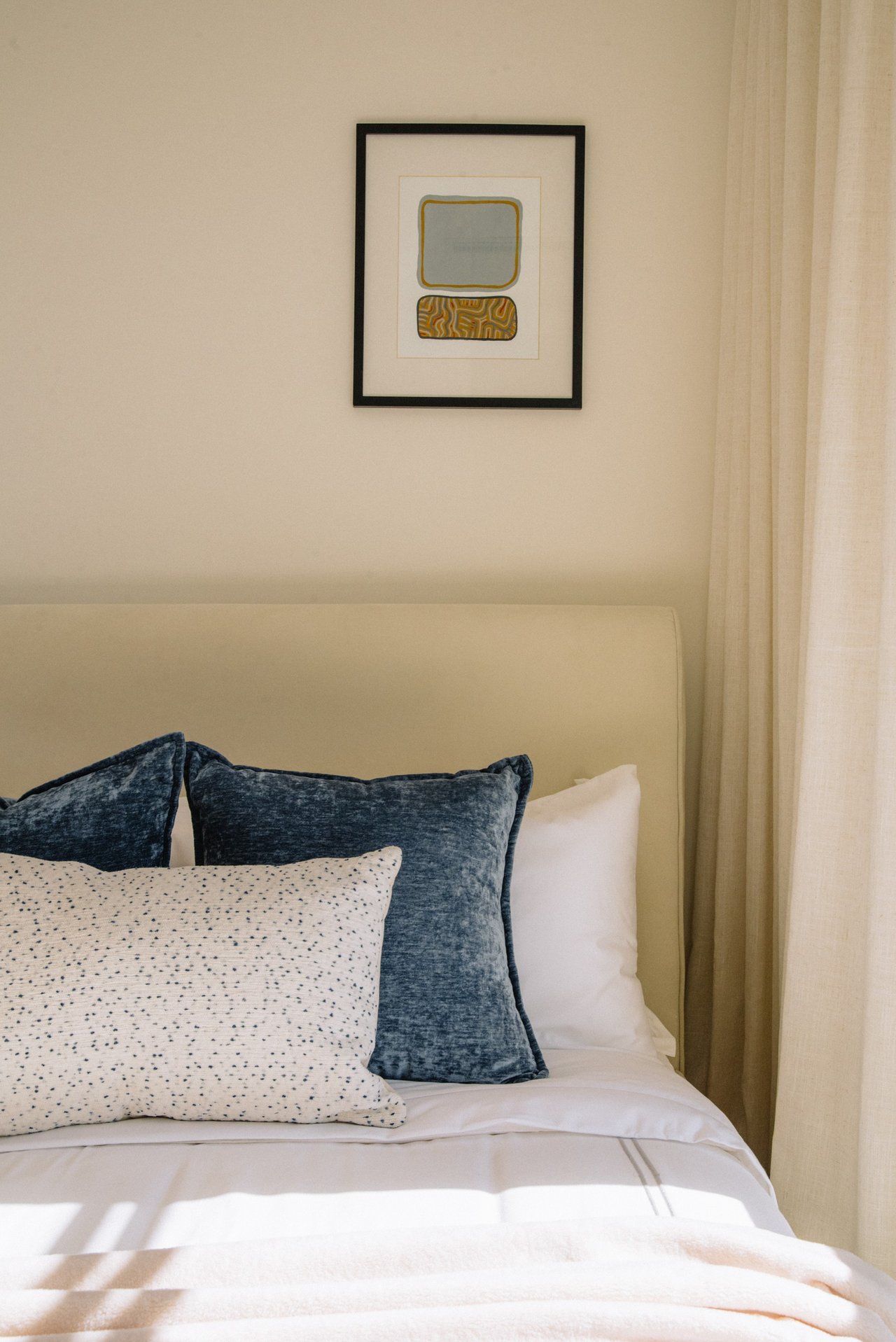 Bedroom with blue and patterned pillows, a cream headboard, framed art, and sheer curtains.