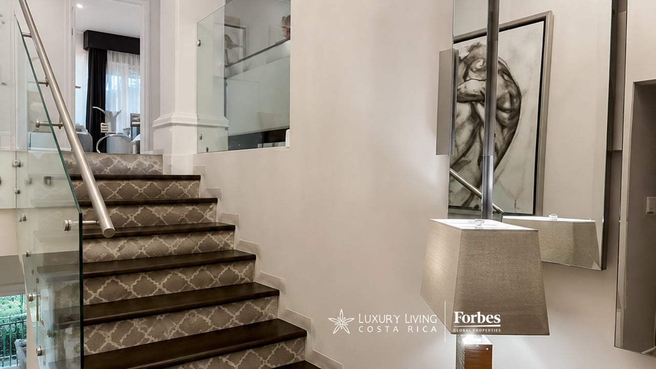 Staircase with dark patterned steps, glass railing, modern floor lamp, and framed abstract artwork.