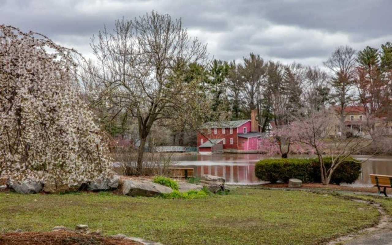 A vibrant red building overlooking a tranquil pond in Demarest, surrounded by blooming spring trees and green grass.