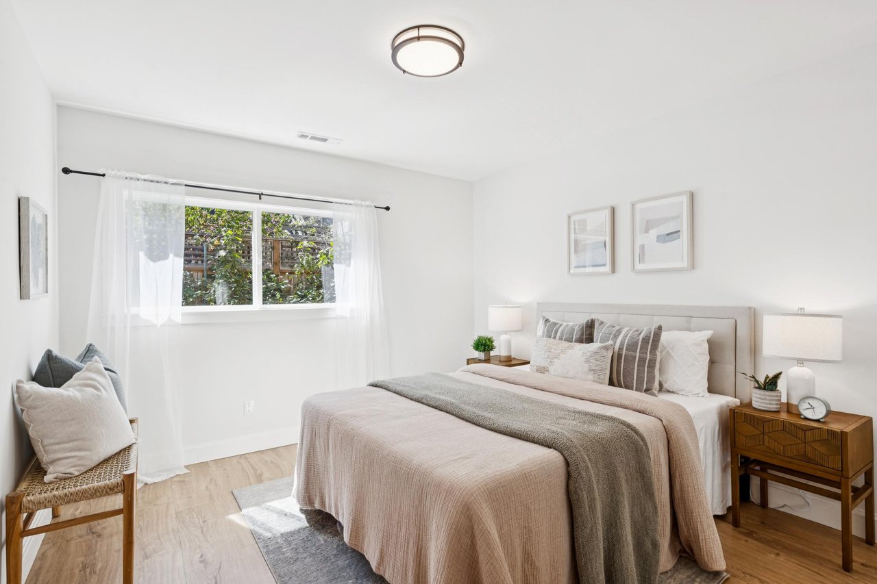 Staged bedroom with a neutral bed, matching bedside tables, and a window overlooking greenery.