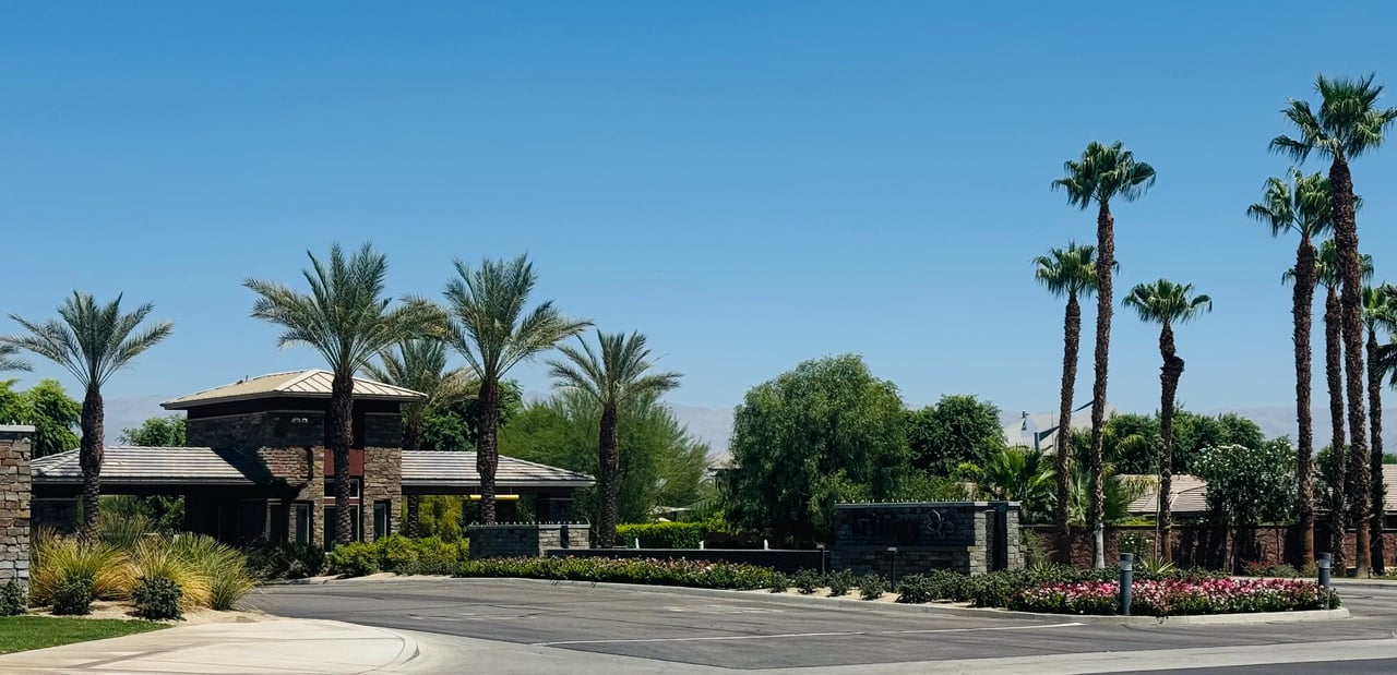Palm-lined residential community entrance with a stone gatehouse and landscaped flower beds.