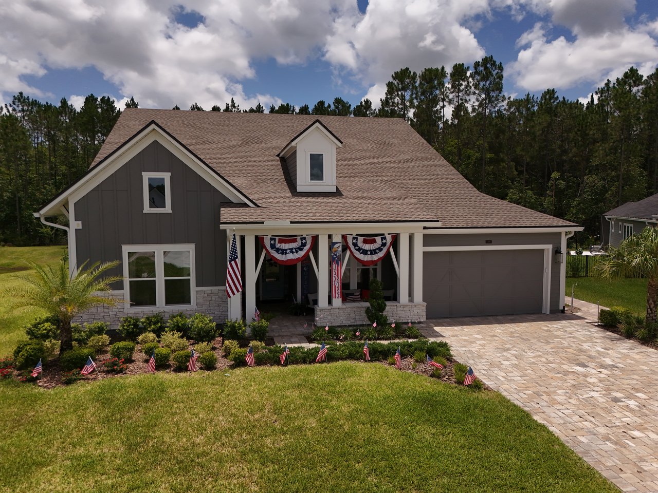 Gray two-story house with patriotic porch bunting, landscaped front yard, and stone-paver driveway.