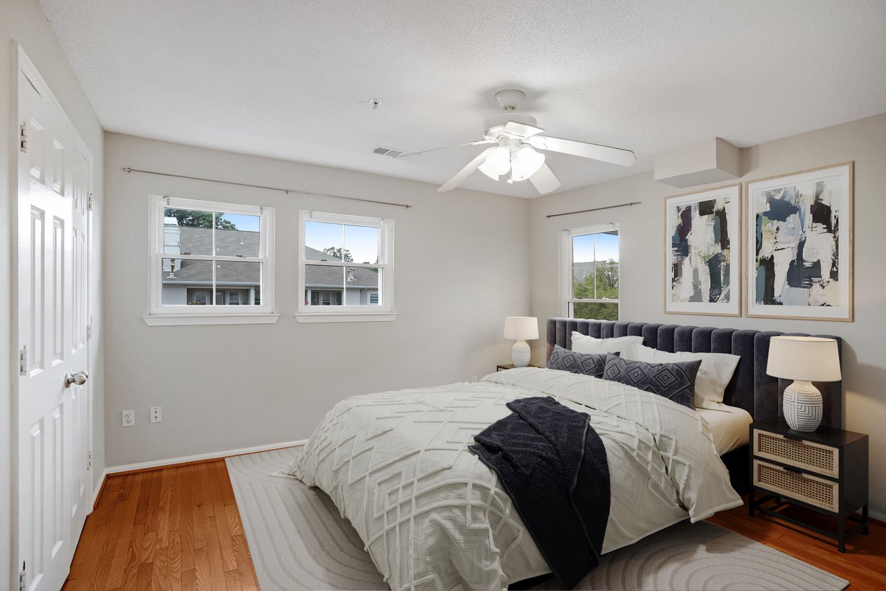 Staged bedroom with a charcoal upholstered headboard, white bedding, three windows, and a ceiling fan.