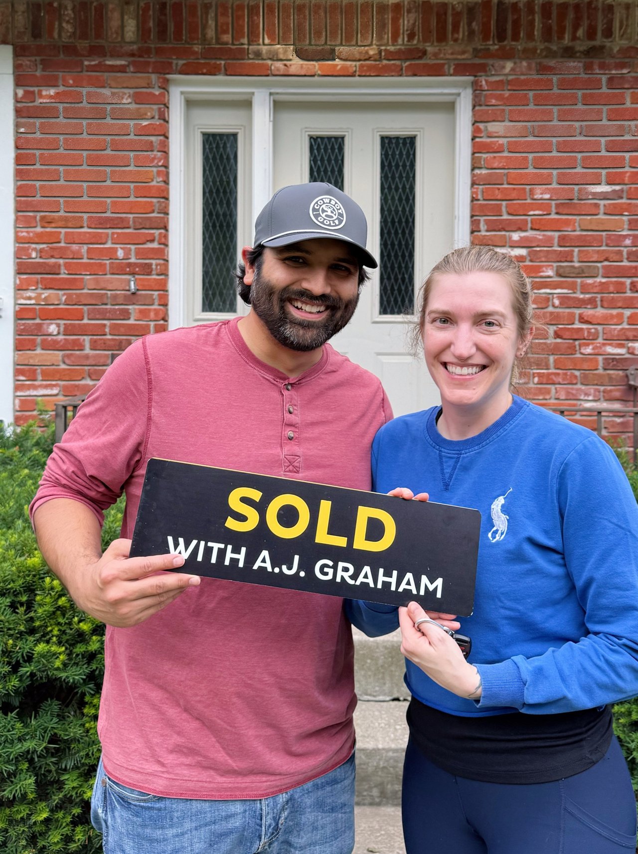 Two smiling adults hold a SOLD WITH A.J. GRAHAM sign outside a brick home.
