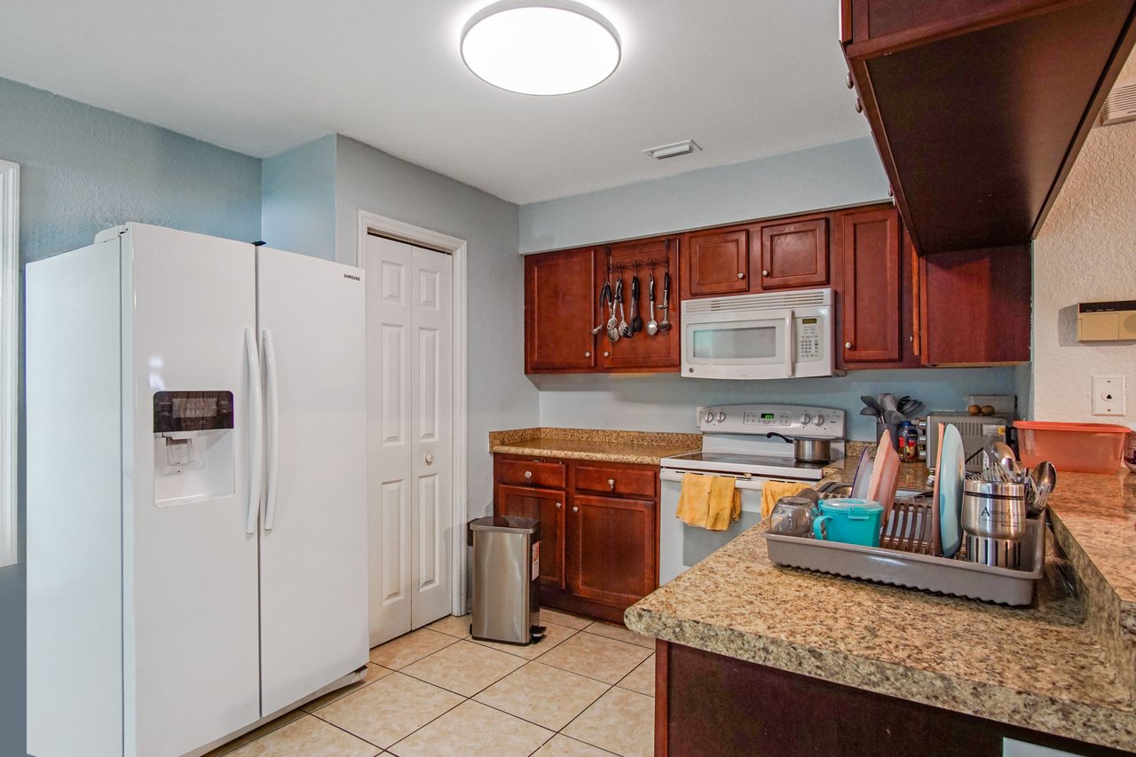 “Kitchen at 2506 Nugget Lane in Tallahassee, Florida, featuring granite countertops, dark wood cabinets, and tile flooring. Appliances include a white side-by-side refrigerator, electric range with overhead microwave, and a stainless steel sink. A pantry door provides additional storage.