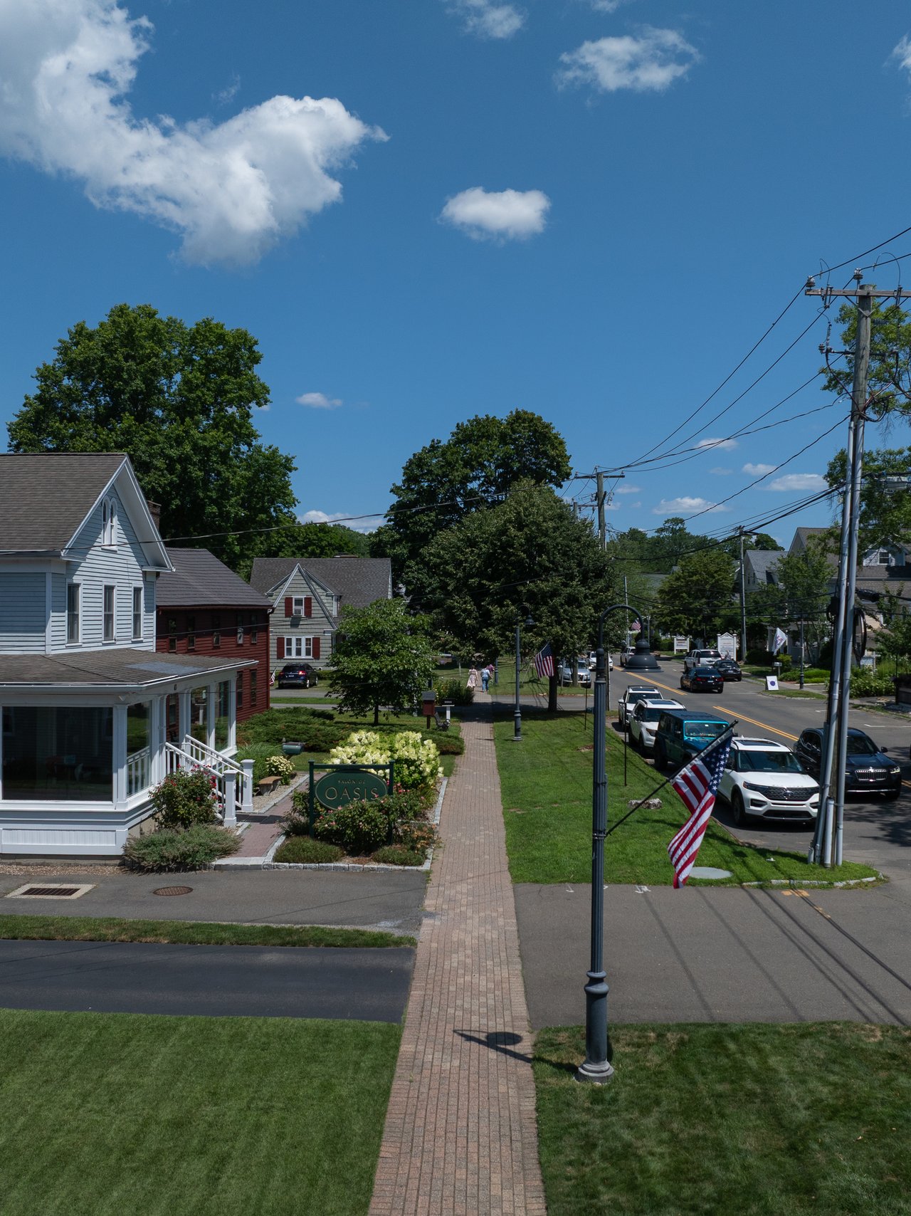 Small-town street with shops, a brick sidewalk, parked cars, and American flags.