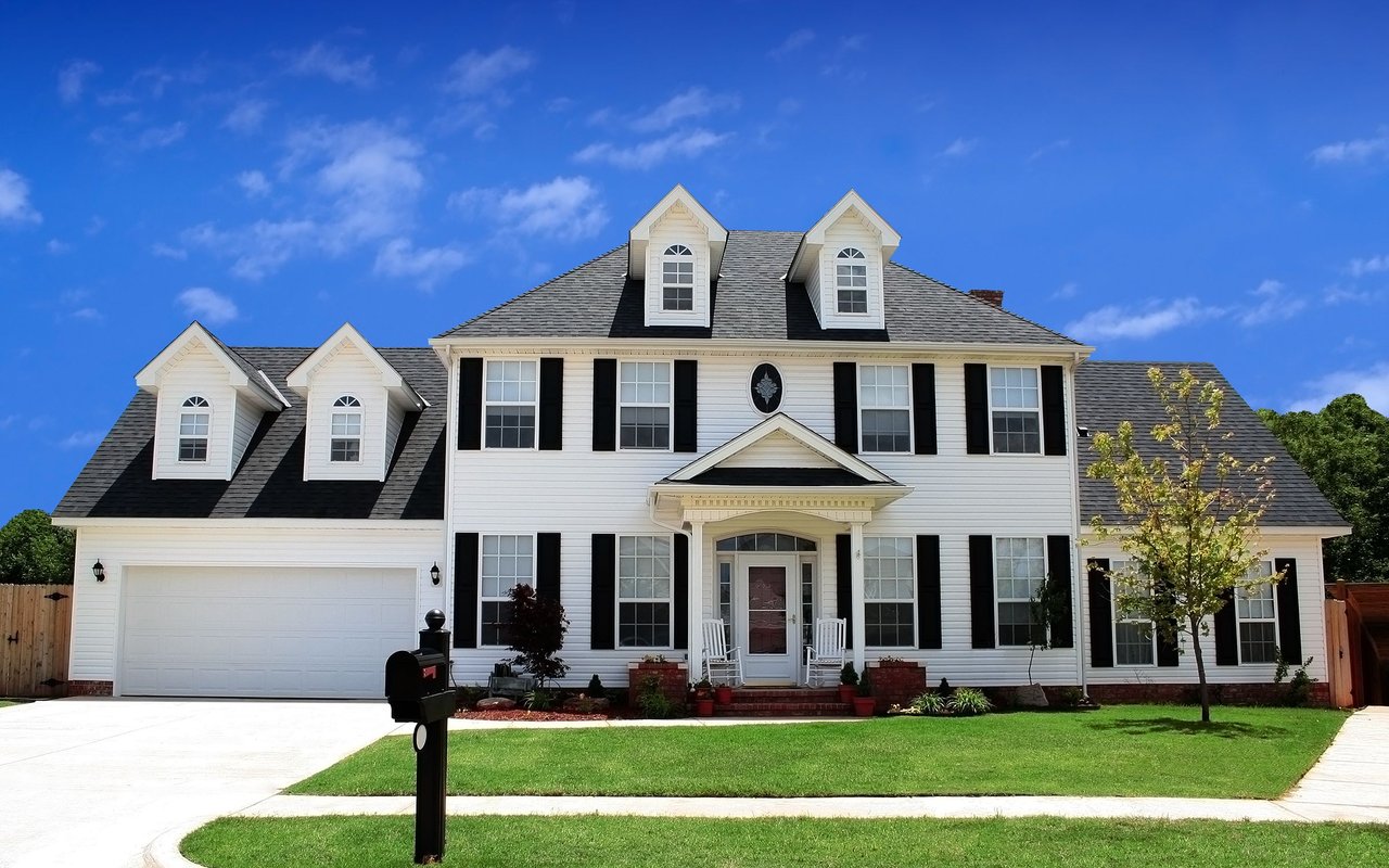 White two-story house with black shutters, multiple dormers, an attached garage, and a broad green lawn.