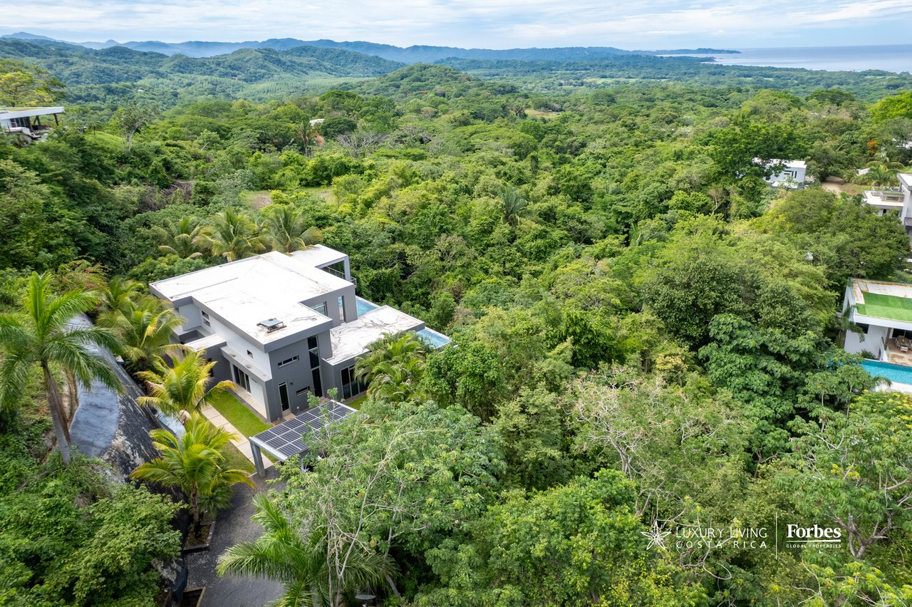 Modern home with solar panels and pool surrounded by tropical forest, with the ocean in the distance.