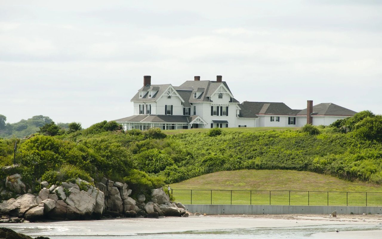 Large white coastal house above a rocky shoreline and fenced lawn.