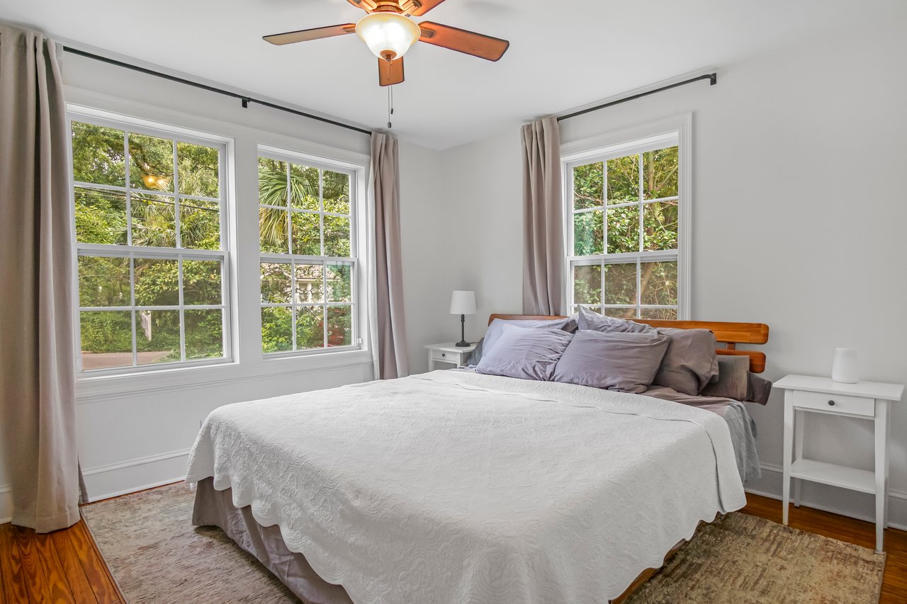 Bright bedroom with a large bed, white bedding, and gray pillows under two windows with curtains. Greenery visible outside, creating a serene atmosphere.