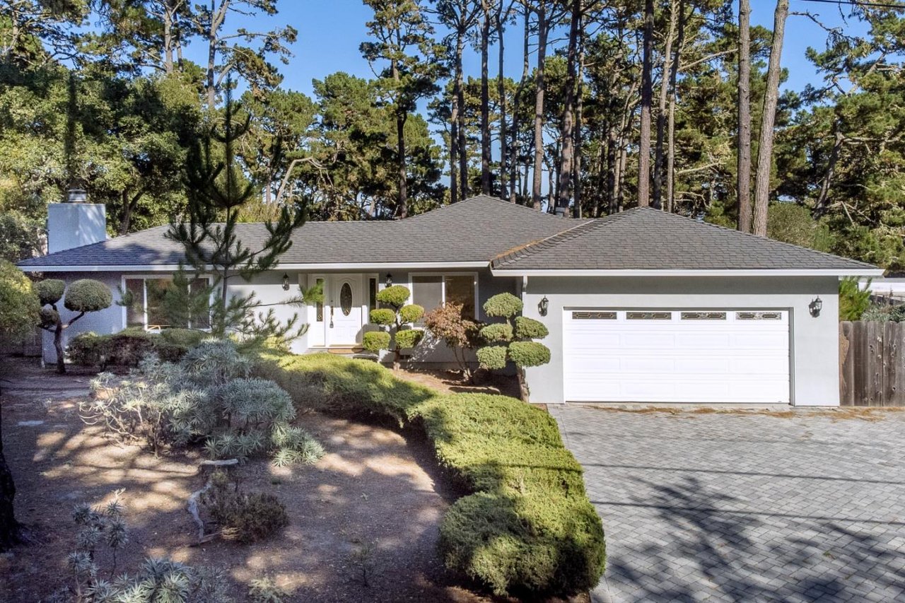 Single-story white home with a two-car garage, landscaped yard, and tall pines behind it.