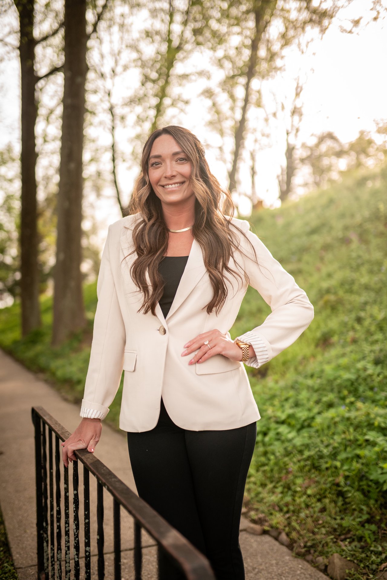 Megan Franc in a white blazer standing beside a railing on a tree-lined path.