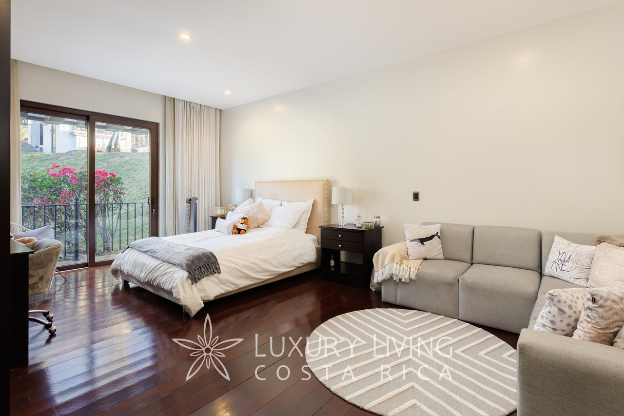 Bedroom with a white bed, gray sofa, and sliding doors overlooking greenery and pink flowers.