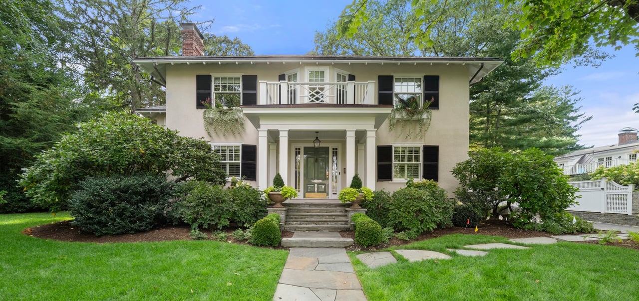 Two-story cream house with black shutters, balcony, columned entry, and landscaped front lawn.