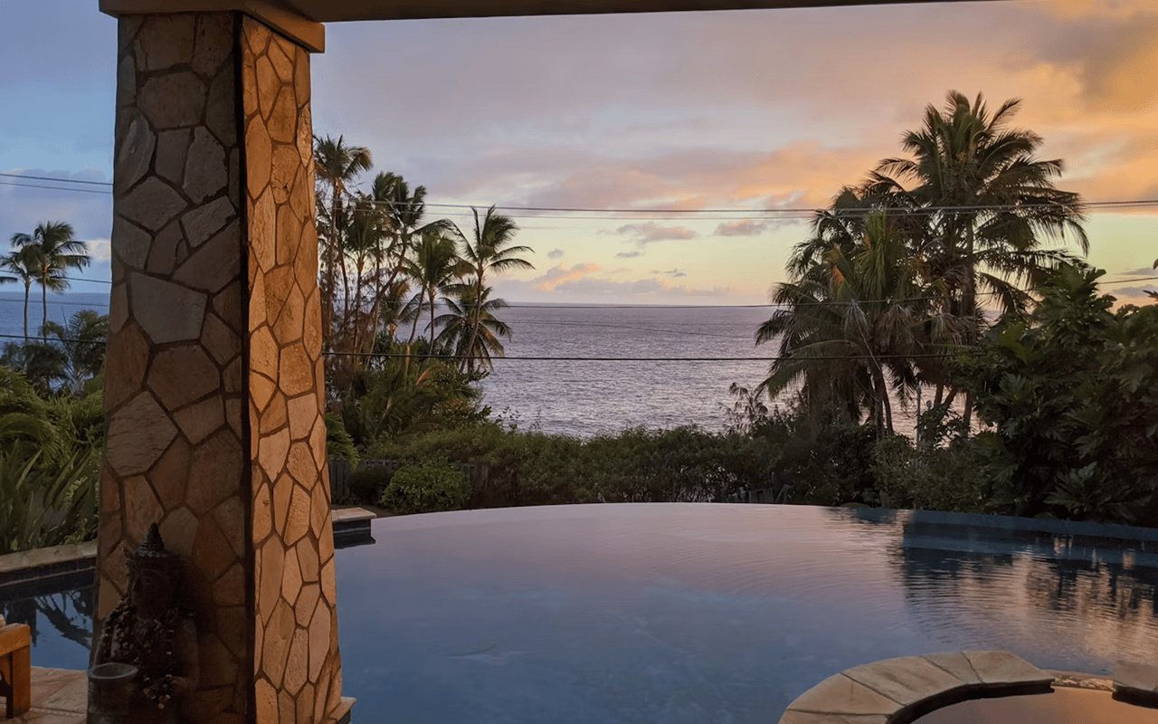 Ocean-view infinity pool framed by palm trees and a stone column at sunset.