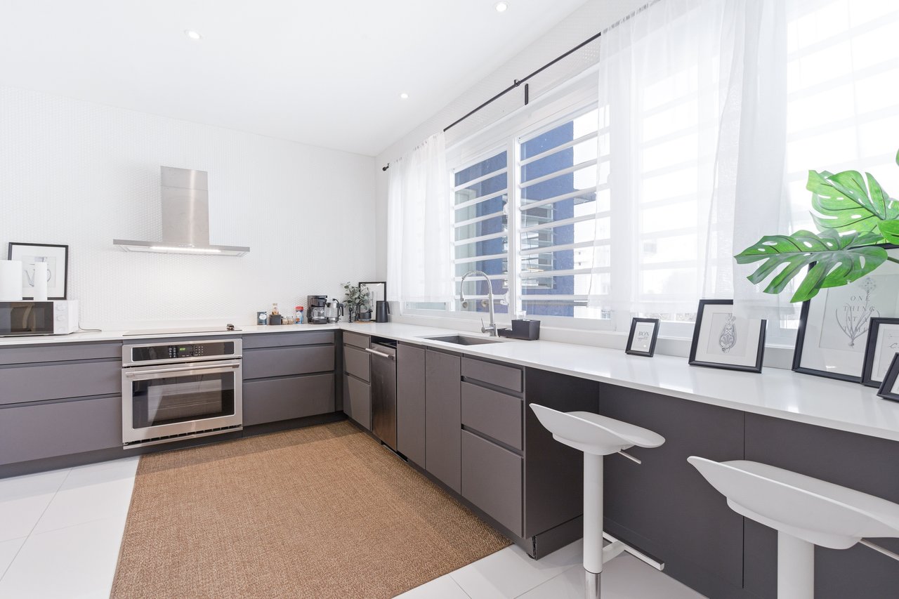 Modern kitchen with dark cabinets, a long white counter, and two stools beside large windows.