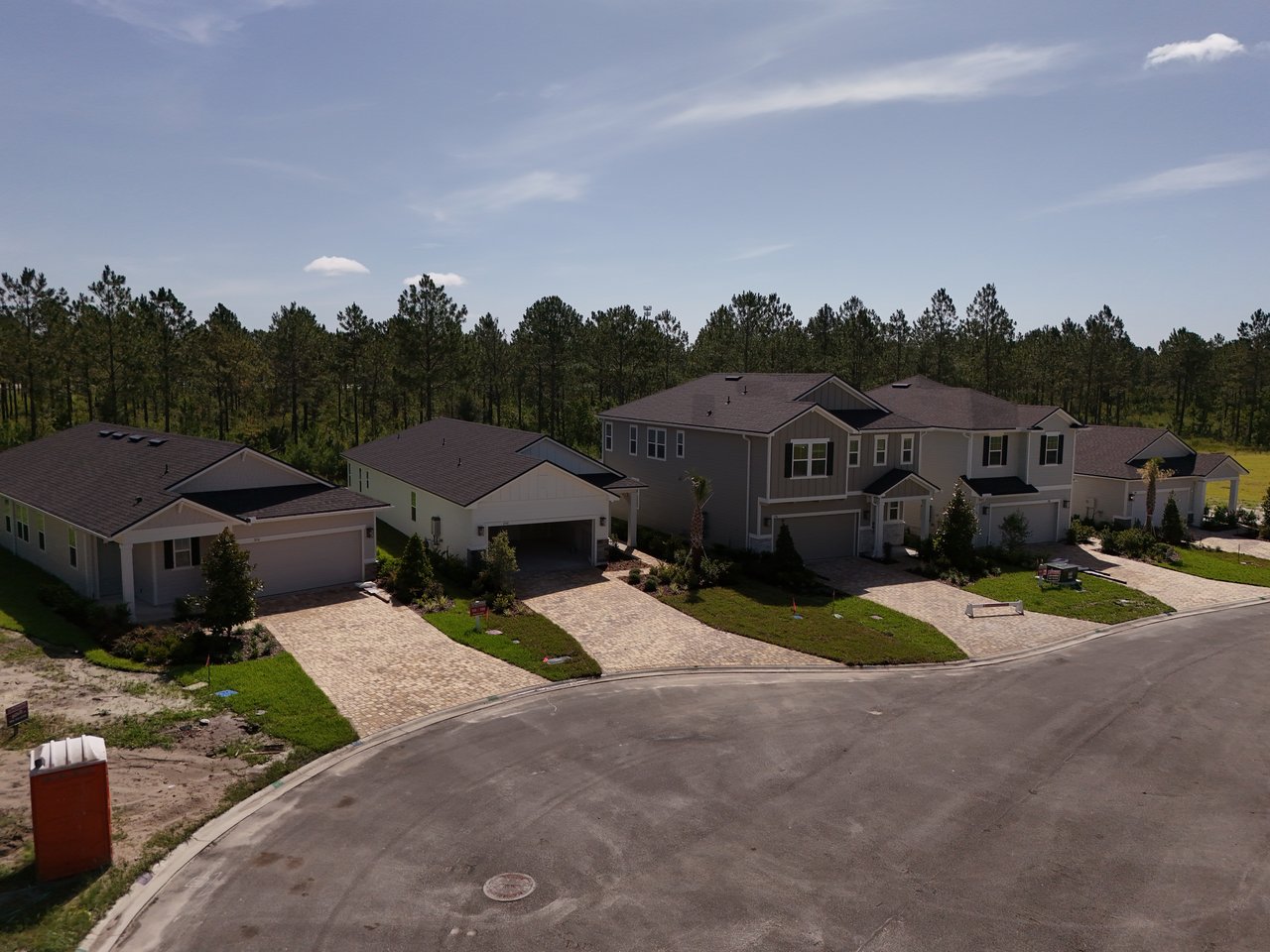 Several newly built homes line a curved street, with a pine tree line behind them.