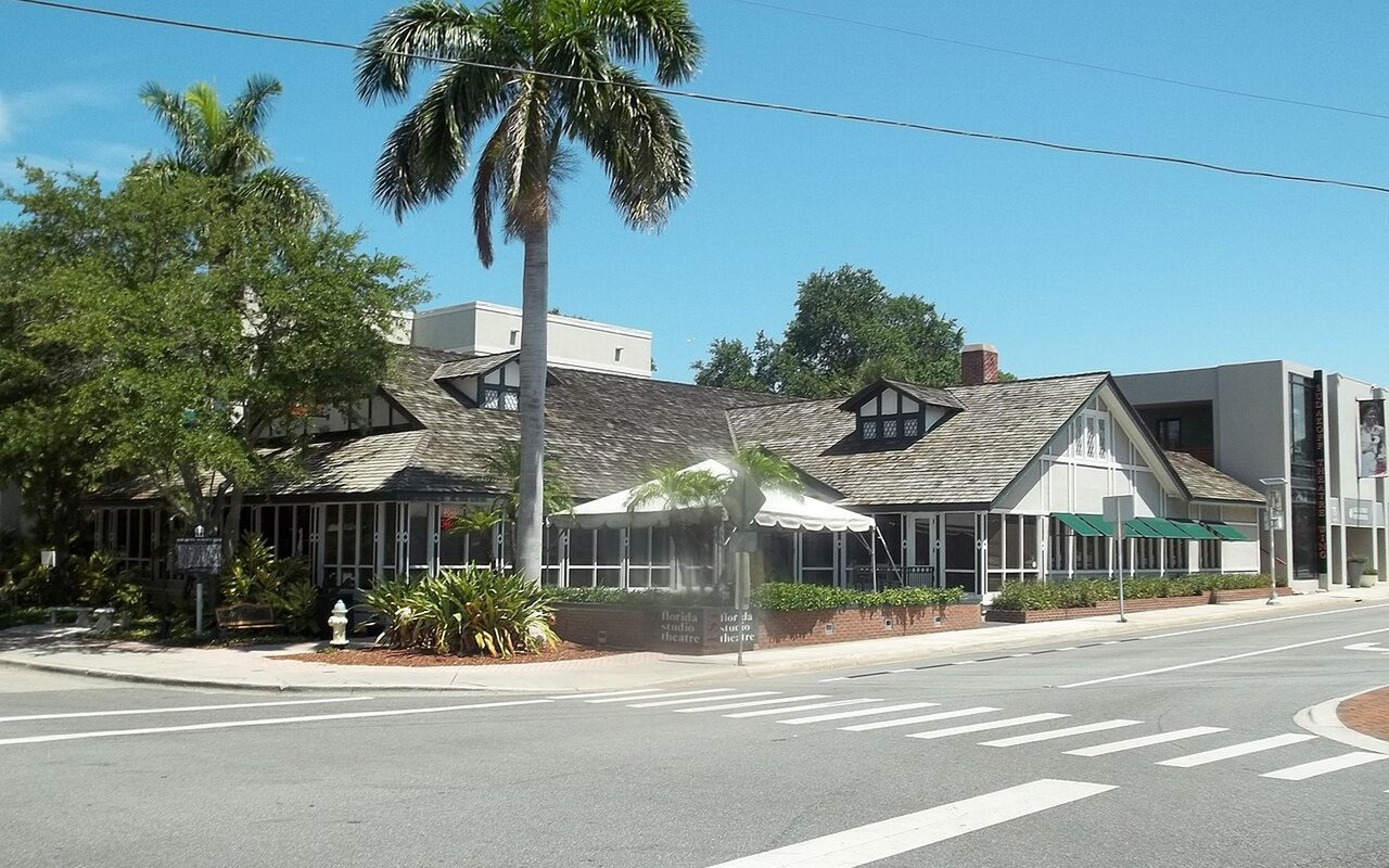 Florida Studio Theatre exterior with shingled gables, green awnings, and palm trees beside a sunny intersection.