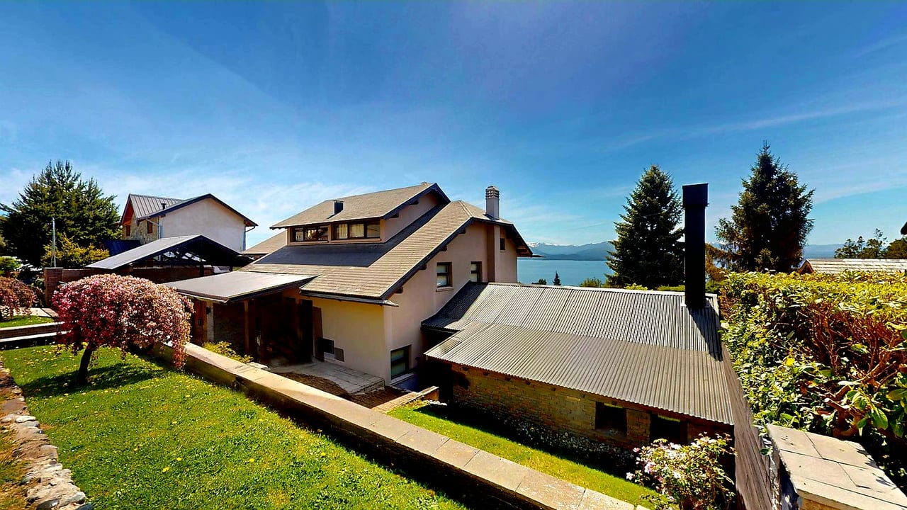 Multi-level house with sloped roofs, landscaped lawn, and a lake and mountains beyond.