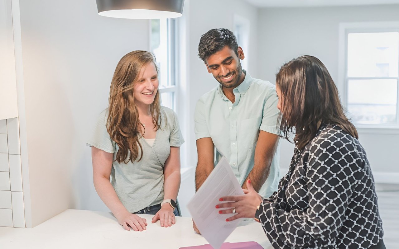 Real estate agent reviewing paperwork with a smiling couple at a kitchen counter.