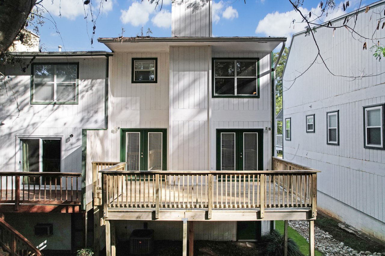 Rear exterior view of the townhome at 529 Collinsford Road, Tallahassee, Florida, featuring white vertical siding with green trim and a spacious elevated wooden deck with railings. French doors with glass panes open to the deck from the main living area, and windows provide natural light to both levels of the home.