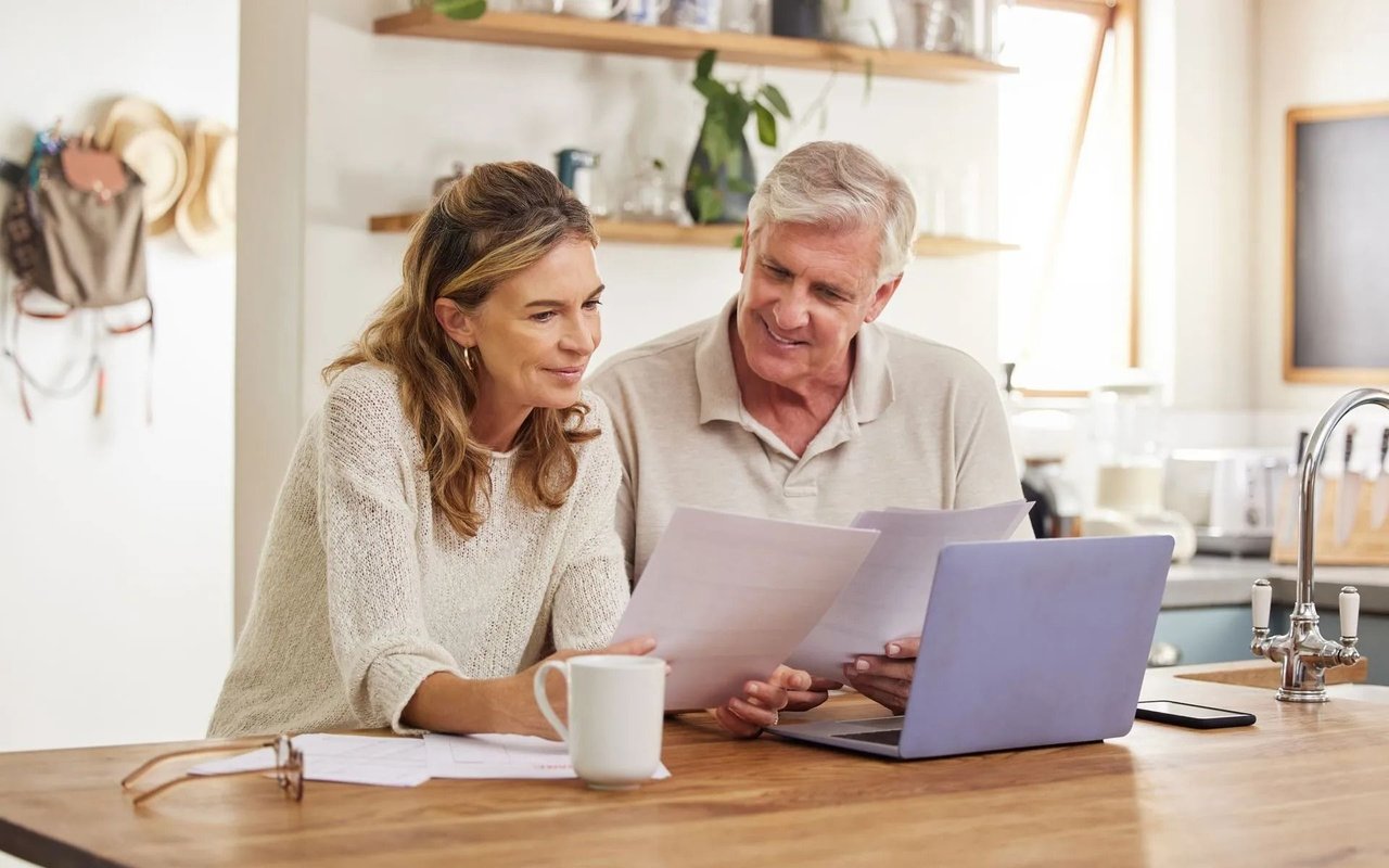 A couple reviews financial papers beside a laptop at a kitchen island.