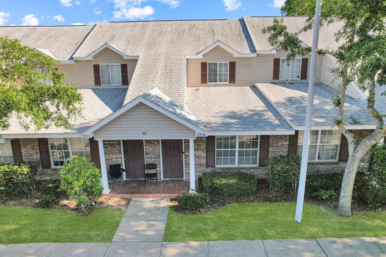 Aerial view of a row of townhouses with light gray roofs and brick exteriors. Lush green lawns and trees frame the entrance, evoking a peaceful suburban scene.