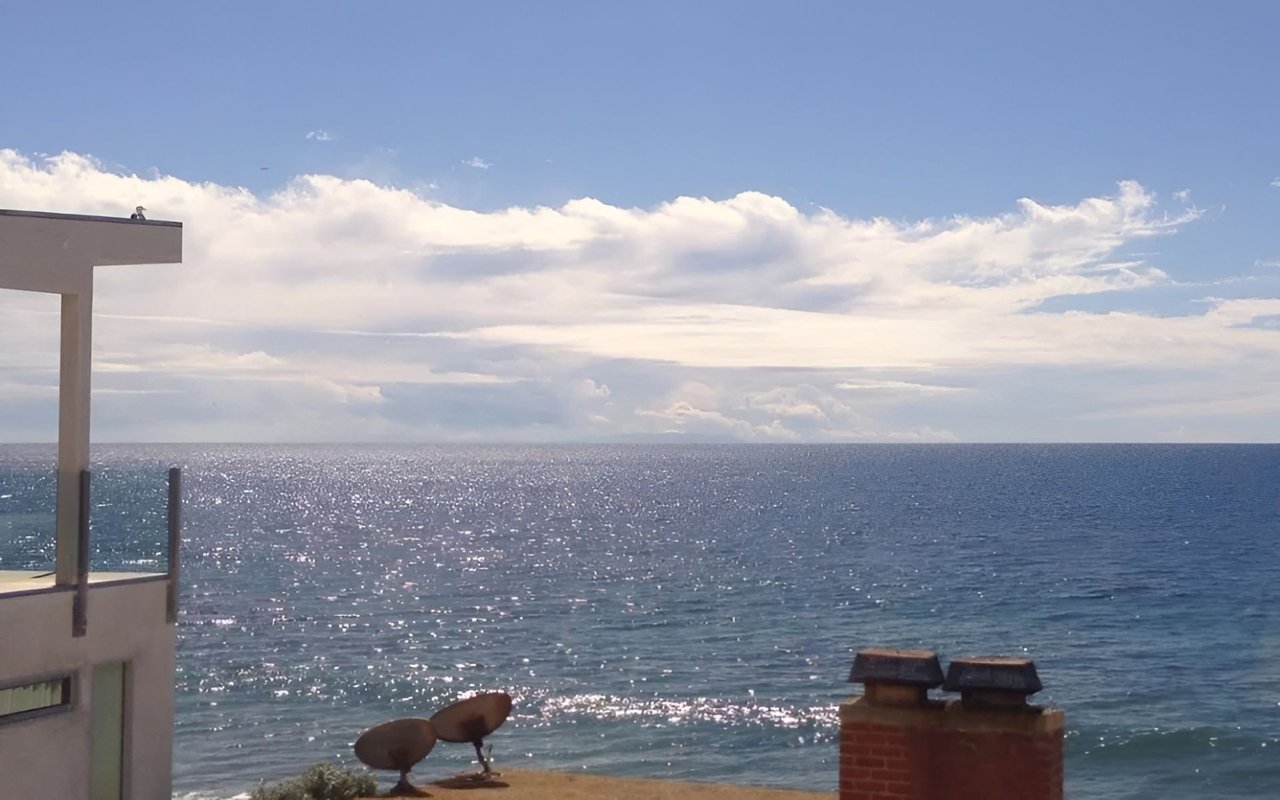 Ocean view from beside a building, with sunlight sparkling on the water beneath scattered clouds.